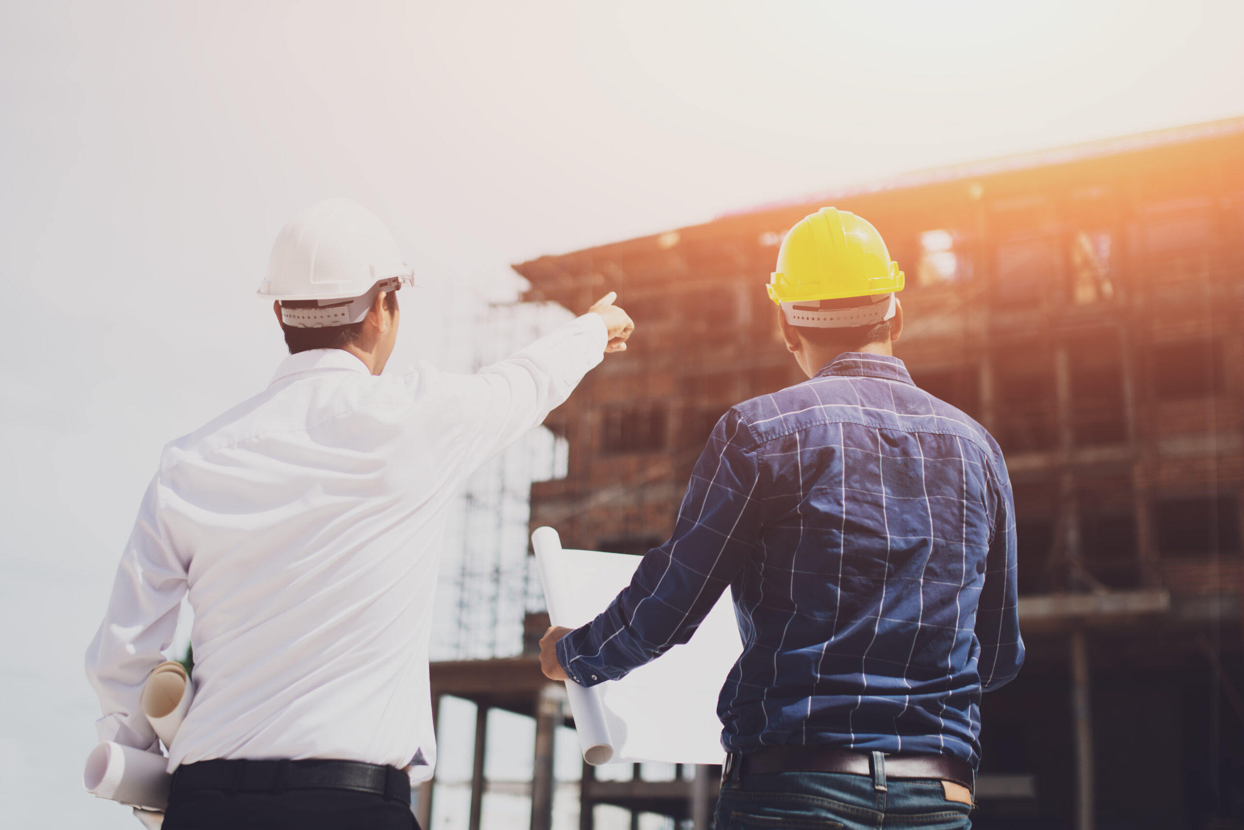two men in hard hats pointing up at a building under construction looking holding plans and conversing